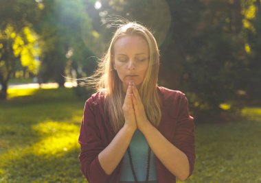 Güneş ışığı altında meditasyon yapan genç bir kadın. Gözleri kapalı, elleri dua pozisyonunda..