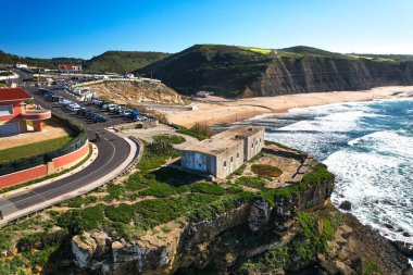 Okyanus dalgaları, kumlu plaj, dik uçurumlar, park alanı ve güneşli bir günde tarihi deniz manzaralı Portekiz kıyısı manzarası. Arkaplanda yeşil tepeler