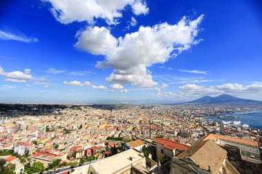 Napoli, İtalya 'nın panoramik manzarası, renkli binalar, arka planda Vesuvius Dağı, ve dramatik bulutlarla dolu parlak mavi bir gökyüzü. Yazın manzaralı kıyı kenti manzarası