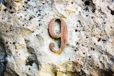 9 numaralı köylü, engebeli bir taş duvara monte edilmiş. Doğal aşınma ve liken izleriyle yaşlanmış metal doku. Adres, işaret, zaman ve çürüme kavramları için ideal
