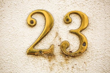 Krem rengi desenli bir duvara 23 numaralı pirinçten ev monte edilmiş. Rakamlar yaş belirtileri gösteriyor. Örümcek ağları ve toprak üste birikiyor. Zaman ve terk edilme hissi ekleniyor.