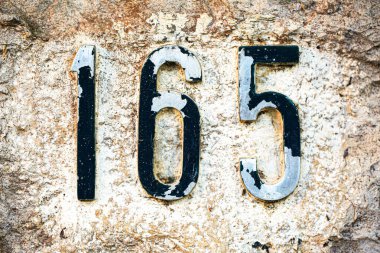 165 numaralı eskimiş ev, siyah, kabuk soyulmuş beyaz boya, kaba, desenli taş bir yüzeye monte edilmiş. Eskimiş görünüş ve yontulmuş bitişi ona kırsal, klasik bir karakter veriyor.