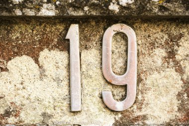 19 numaralı metal ev yıpranmış taş bir duvara monte edilmiş. Görünür pas ve doğal renk bozulması var. Rakamlar kalın ve biraz yıpranmış, kırsal ve eskimiş mimari bir cazibe katıyor.