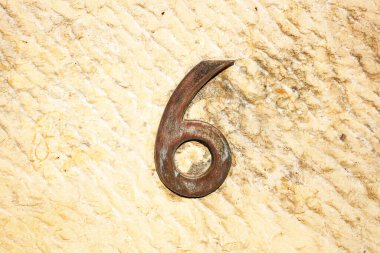 Kaba, sarımsı taştan bir duvara monte edilmiş 6 numaralı kırsal metal. Rakamlar yaşlanmanın belirtilerini gösteriyor. Deri kaplamalı doğal taş arka planıyla güzel tezat oluşturuyor.