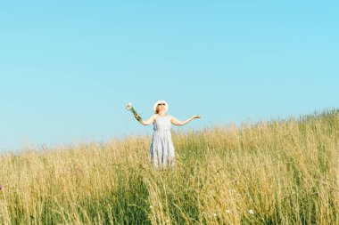 Parlak bir yaz gününde, uzun bir çim tarlasında kır çiçekleriyle mutlu olgun bir kadın..