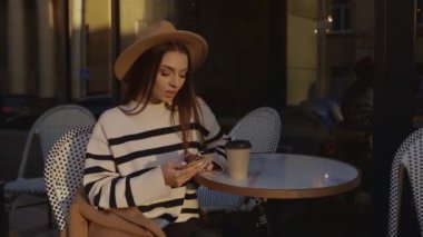 Şapkalı güzel bir genç kız bir sokak kafesinde oturuyor ve sosyal ağlarda gezinmek ve mesajlar göndermek için cep telefonu kullanıyor.