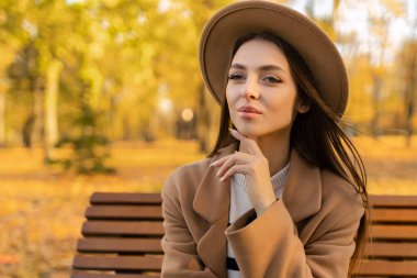 Parkta yürüyen, yakışıklı, uzun saçlı, güler yüzlü bir kadının portresi. Kahverengi ceketli, sonbahar modasına uygun, sokak tarzı şapkalı..