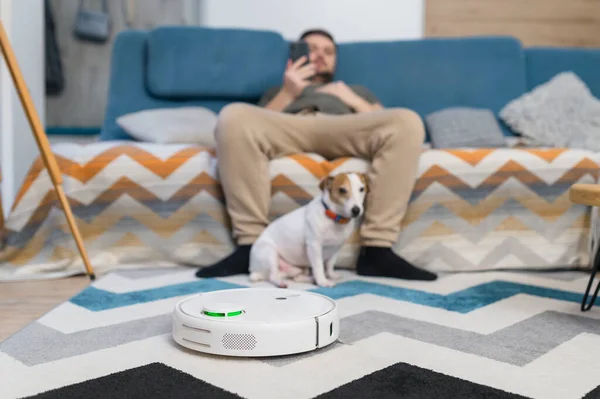 Man couple sitting on sofa with dogs cleaning floor using vacuum robot at home.
