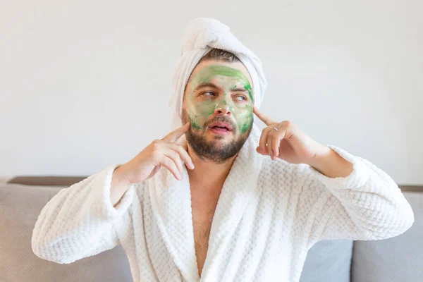 stock image Surprised guy spends hours treating and caring for his skin, applies green face mask to pamper himself, wearing a soft bathrobe and a towel on his head. 