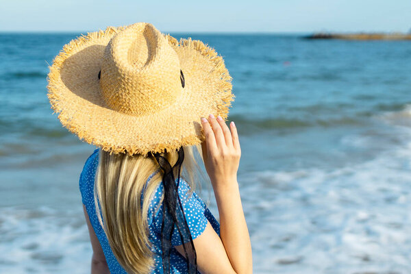 Woman in dress with straw hat walking by sea on sunny day, back view. 