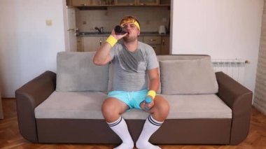 Spor kıyafetli ve ıslak tişörtlü komik bir adam koltukta dambıllarla oturur ve bira içer.. 