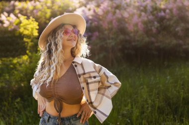 Şapkalı ve güneş gözlüklü seksi kıvırcık sarışın çiçek açan ağaçların yanında poz veriyor..