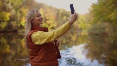 Mutlu, sarışın bir kadın güler yüzlü, konuşur internette konuşur sonbahar parkında tek başına cep telefonu teknolojisi kullanarak selfie çeker.. 