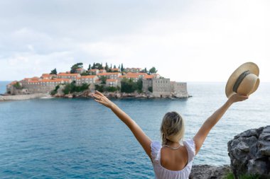 Şapkalı kız turist Karadağ 'ın Sveti Stefan adasında ve denizde tepeden bakıyor.