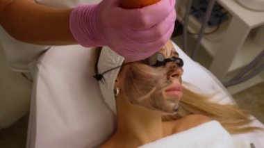 Güzellik uzmanları, güzellik salonundaki bir kanepede uzanan gözlüklü güzel bir müşteriye karbon soyuyorlar. Yüz temizleme konsepti. Kozmetik Hizmetleri. 