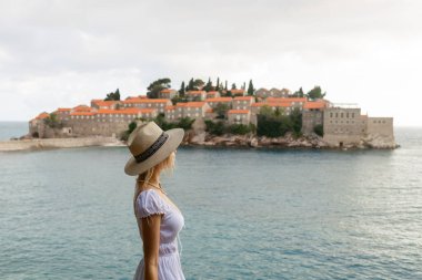 Şapkalı kız turist Karadağ 'ın Sveti Stefan adasında ve denizde tepeden bakıyor.