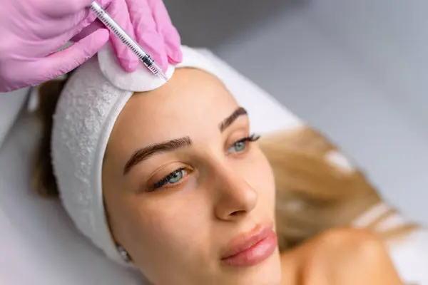 stock image Close up of hands of young cosmetologist injecting botox in female face. She is standing and smiling. The woman is closed her eyes with relaxation.