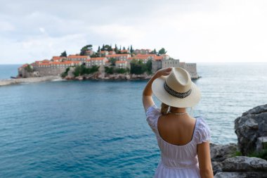 Şapkalı kız turist Karadağ 'ın Sveti Stefan adasında ve denizde tepeden bakıyor.