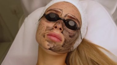 Güzellik uzmanları, güzellik salonundaki bir kanepede uzanan gözlüklü güzel bir müşteriye karbon soyuyorlar. Yüz temizleme konsepti. Kozmetik Hizmetleri. 