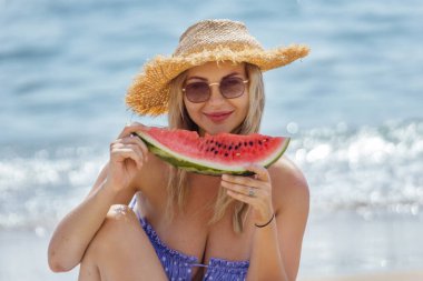 Yüzünde güneş gözlüğü ve şapkası olan, kumsalda karpuz dilimiyle mutlu bir şekilde oturan güzel, sarışın bir kadının portresi..