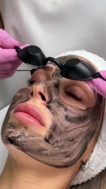 Güzellik uzmanları, güzellik salonundaki bir kanepede uzanan gözlüklü güzel bir müşteriye karbon soyuyorlar. Yüz temizleme konsepti. Kozmetik Hizmetleri. 