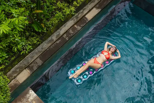 Güneşlenmenin tadını çıkarıyorum. Tropik tatil konsepti. Yüzme havuzunda palmiye ağaçlarıyla şişme yatakta genç bir kadın.
