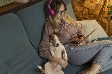 Kanepede bir kız dizüstü bilgisayarında bir köpekle uzanıyor ve kulaklıkla müzik dinliyor. İnternet üzerinden serbest çalışma ve iletişim. 