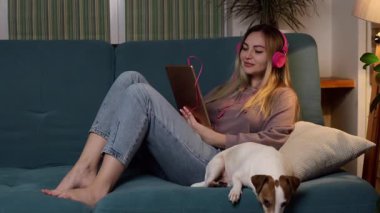 Kanepede bir kız dizüstü bilgisayarında bir köpekle uzanıyor ve kulaklıkla müzik dinliyor. İnternet üzerinden serbest çalışma ve iletişim. 