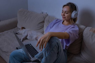 Akşamları evde dizüstü bilgisayarda film izleyen, kulaklık takan ve köpeğiyle kanepede dinlenen genç bir kadın. İçerideki samimi dijital eğlence anı