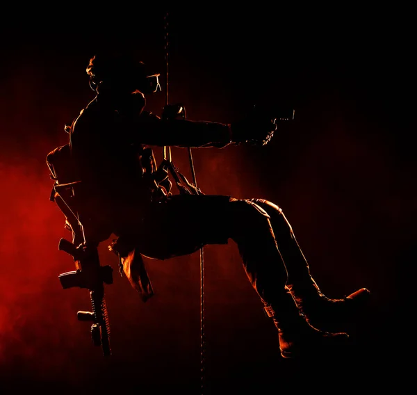 Silhouette of police officer in tactical gear descending from a height, rope exercises with weapons. Tactical rappelling, anti-terror or counter terrorism operation in darkness in rappelling harness