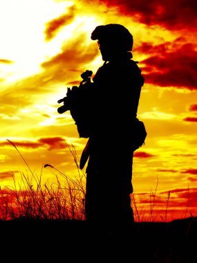 Gün batımında tüfekli Amerikan askerinin silueti arka planda güneş batarken ayakta duruyor.