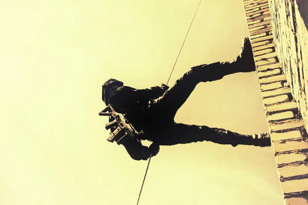 高層ビルから降下する戦術装備の警察官のシルエット 武器でロープ演習 ラッピングハーネスにおける戦術的なラッピング テロ対策 テロ対策 ロイヤリティフリーのストック画像