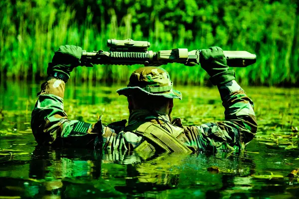 stock image A camouflaged soldier walks through a swamp, submerging himself, his arms and rifle visible, shot from behind, realities of harsh military service