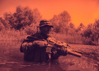 Kamuflaj elbisesi ve şapkası olan bir asker elinde bir tüfekle arka planda ağaçlarla birlikte turuncu renkli bir gökyüzünün altında bataklıkta yürüyor..