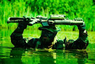 Suda kamufle olmuş asker kafasının üstünde bir tüfek tutarken yemyeşil sularda kısmen daldırılmış..