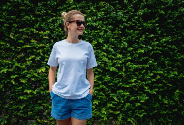 Yaprak duvarın arka planında beyaz, boş tişört giyen bir kadın model.