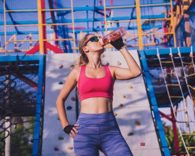 Sporcu genç bir kadın idman yaptıktan sonra su içiyor ve eğitim kampındaki yapay kayaya tırmanıyor.