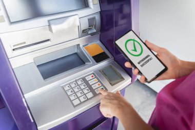 Akıllı telefonu dikkatsizce ATM 'nin yakınındaki parayı çekmek için kullanan genç bir kadın..