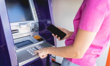 Akıllı telefonu dikkatsizce ATM 'nin yakınındaki parayı çekmek için kullanan genç bir kadın..