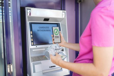 Akıllı telefonu dikkatsizce ATM 'nin yakınındaki parayı çekmek için kullanan genç bir kadın..