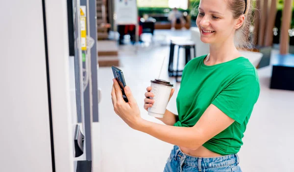 Young woman paying for coffee at vending machine using contactless method of payment.
