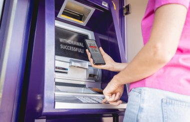 Akıllı telefonu dikkatsizce ATM 'nin yakınındaki parayı çekmek için kullanan genç bir kadın..