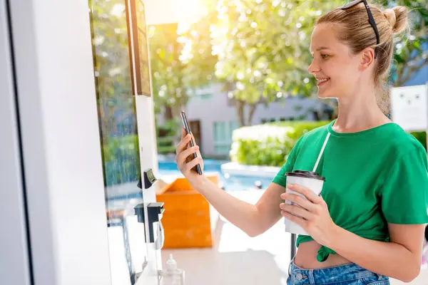Young woman paying for coffee at vending machine using contactless method of payment.
