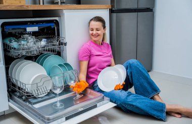 Genç bir kadın bulaşık makinesinin yanında yerde oturuyor..