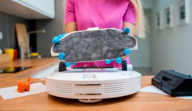 Genç bir kadın temizlik yaptıktan sonra robot elektrik süpürgesini pislikten temizliyor.