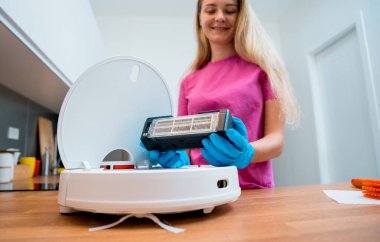 Genç bir kadın temizlik yaptıktan sonra robot elektrik süpürgesini pislikten temizliyor.