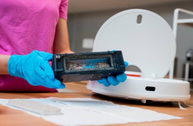 Genç bir kadın temizlik yaptıktan sonra robot elektrik süpürgesini pislikten temizliyor.