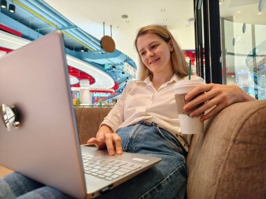 Genç bir kadın canlı bir kafede dizüstü bilgisayarıyla çalışırken kahveden zevk alır.