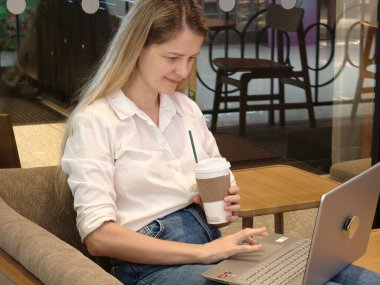 Genç bir kadın canlı bir kafede dizüstü bilgisayarıyla çalışırken kahveden zevk alır.