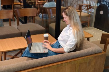 Genç bir kadın canlı bir kafede dizüstü bilgisayarıyla çalışırken kahveden zevk alır.